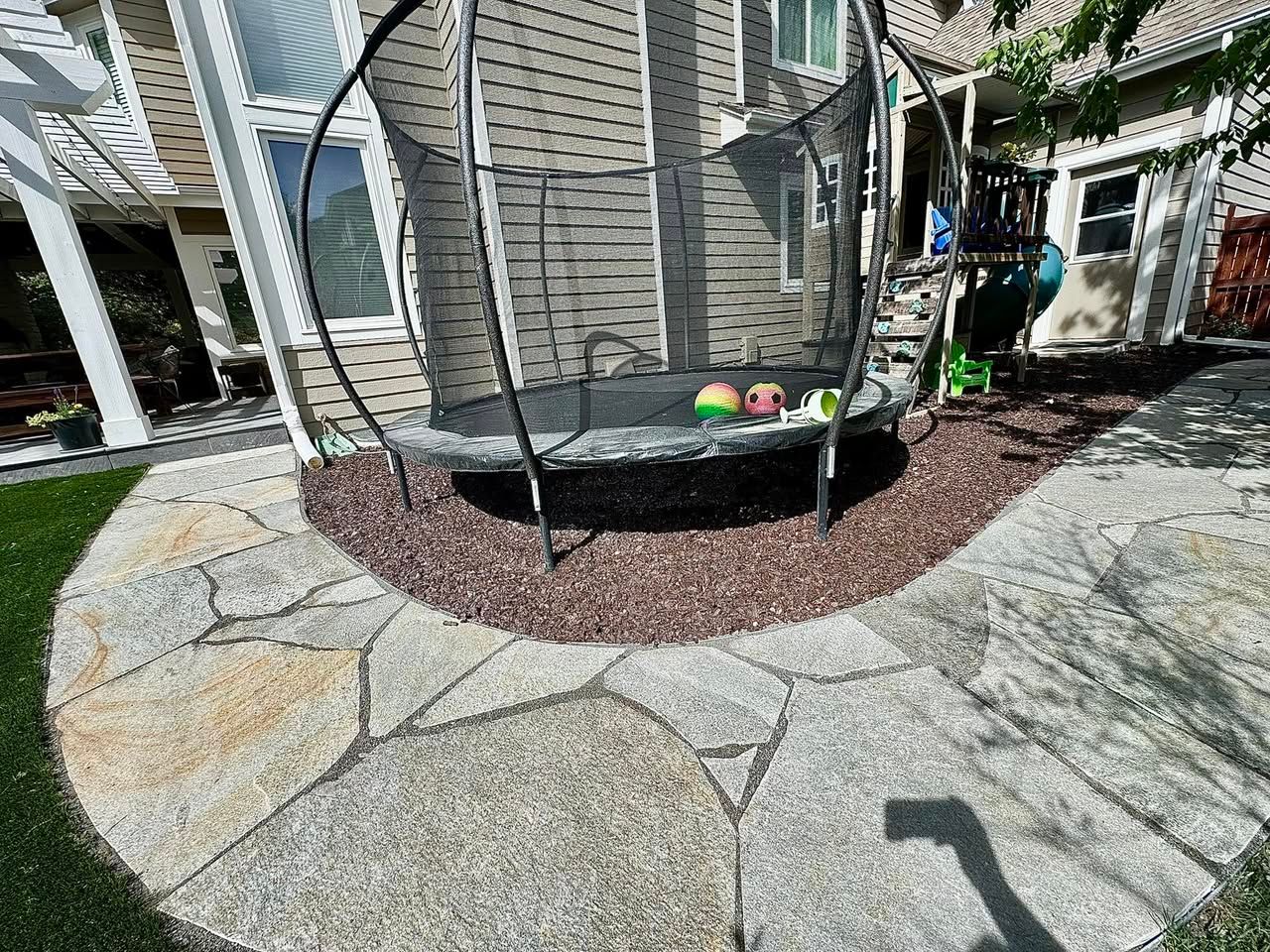 A trampoline sits on reddish-brown mulch surrounded by flagstone pavers and green turf in a backyard.
