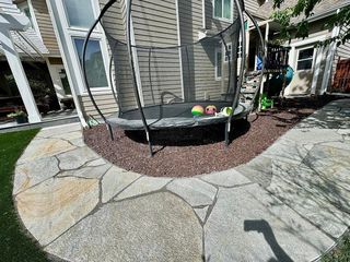 A trampoline sits on a bed of reddish-brown rocks, surrounded by flagstone pavers and green grass.