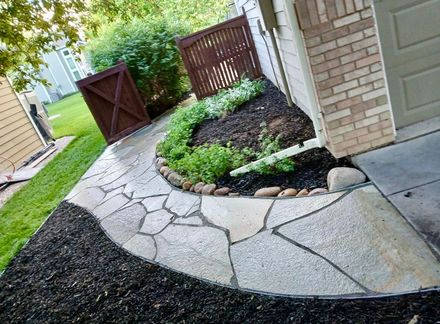 Stone pathway curving around a landscaped flower bed with dark mulch, next to a garage.