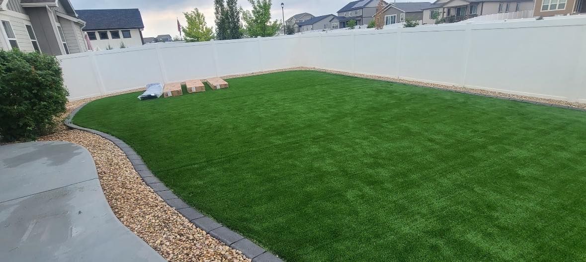 A backyard with green grass, a white fence, and a border of tan rocks.