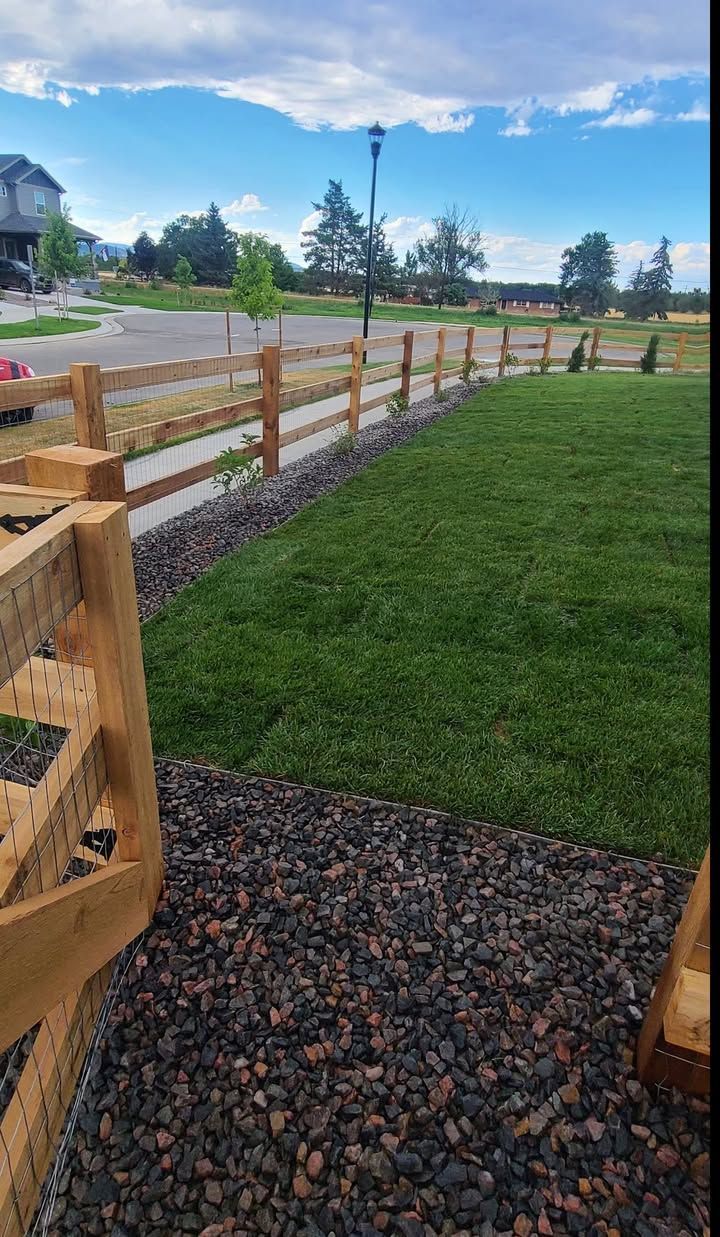 Wooden split-rail fence alongside a green lawn and gravel pathway, under a blue sky.