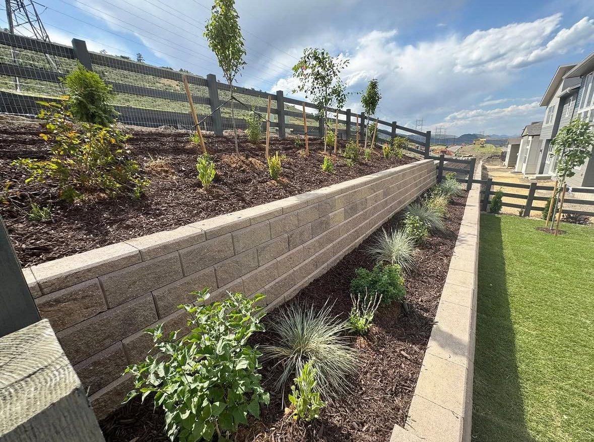 Brick retaining wall with plants, mulch, and wooden fence on hillside under cloudy sky.
