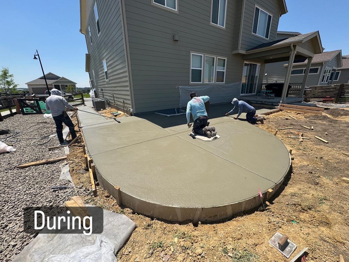 Concrete patio being poured, workers smoothing surface. House exterior in background. Sunny outdoor setting.