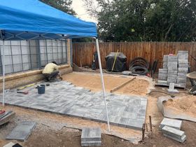 A person laying gray pavers for a patio construction project outdoors under a blue canopy.