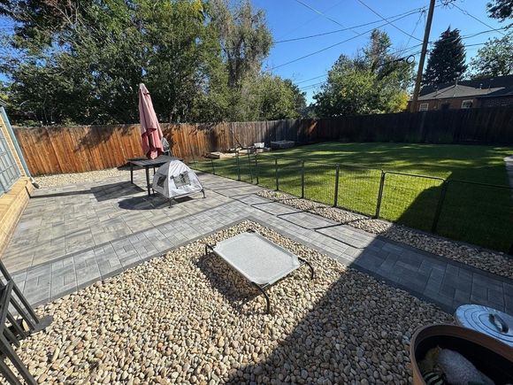 Patio with pavers and gravel leads to a grassy yard bordered by a wooden fence, under a blue sky.