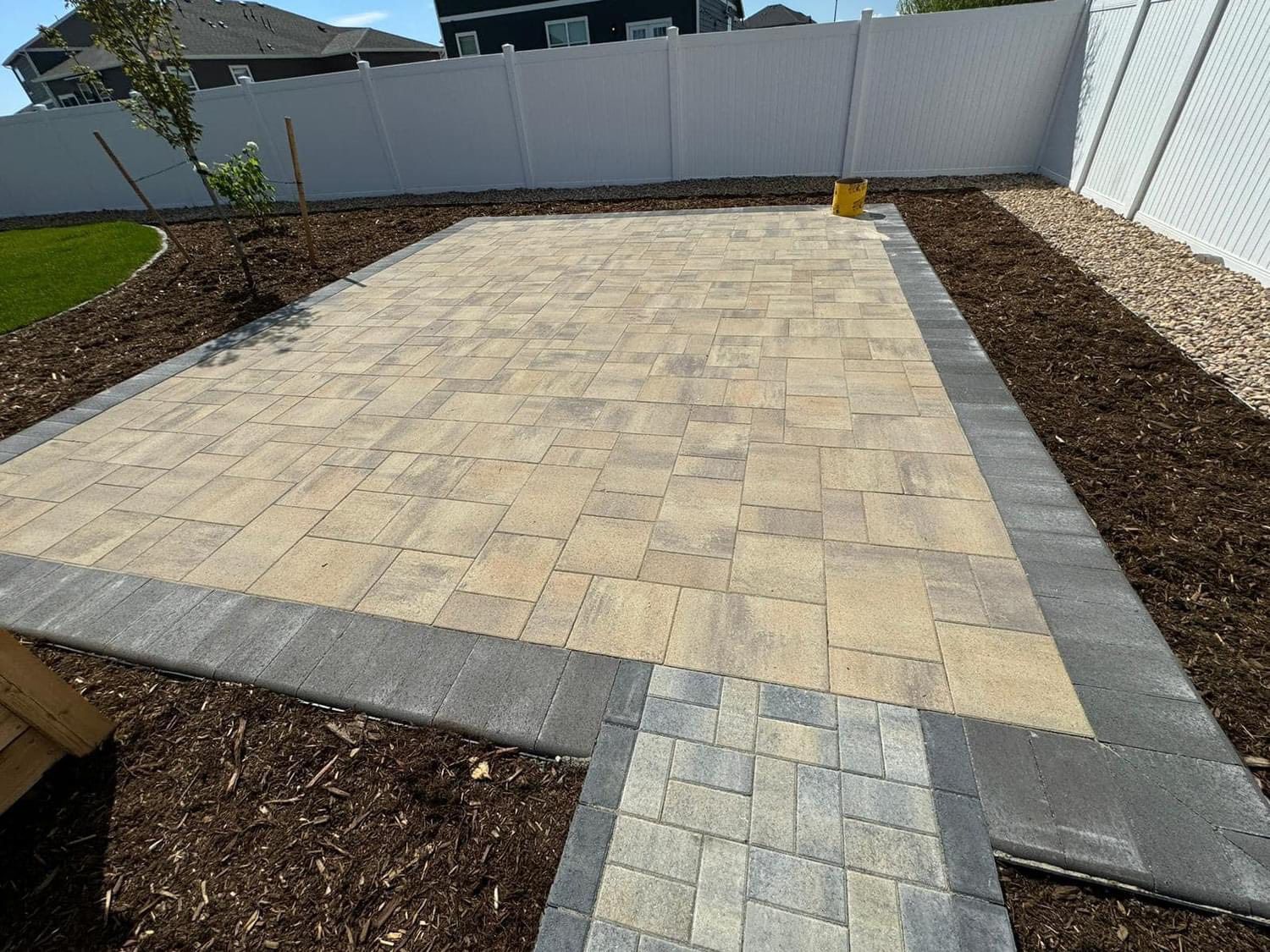 Paver patio in backyard, bordered by dark gray blocks and mulch. White fence in background.