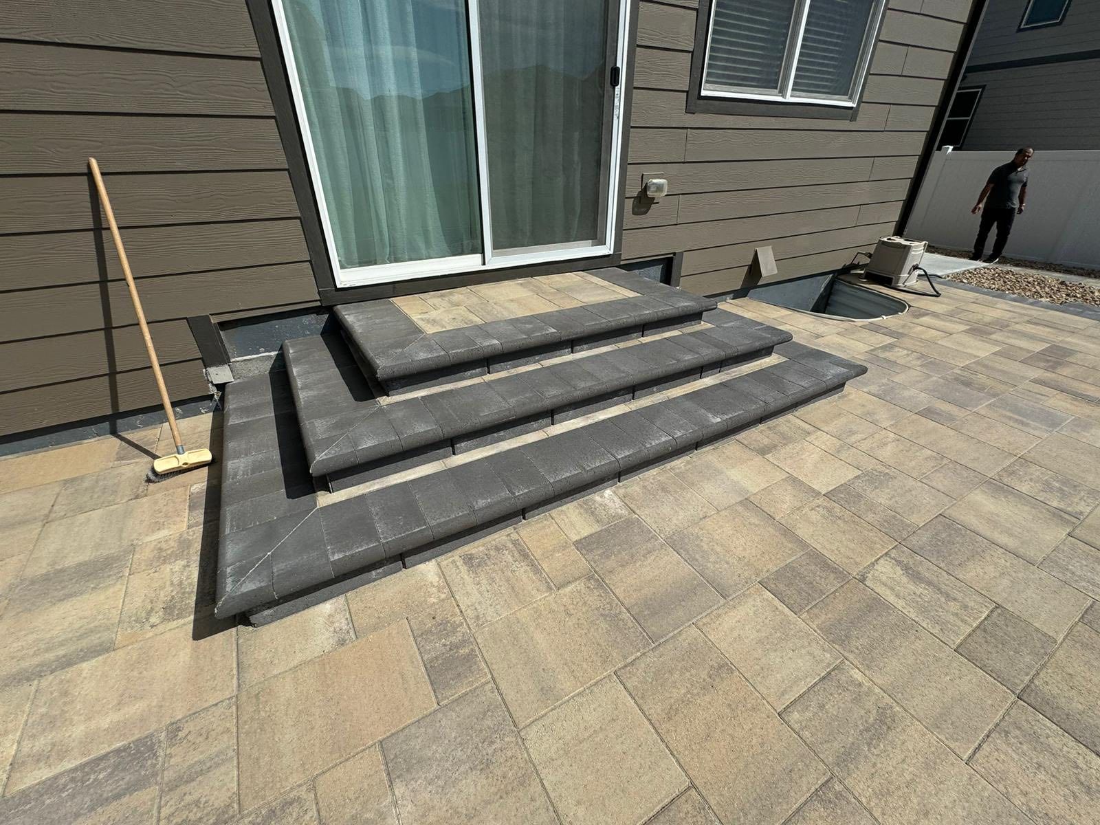 Three-step concrete stairs leading to a sliding glass door, set on a stone patio. A person stands nearby.