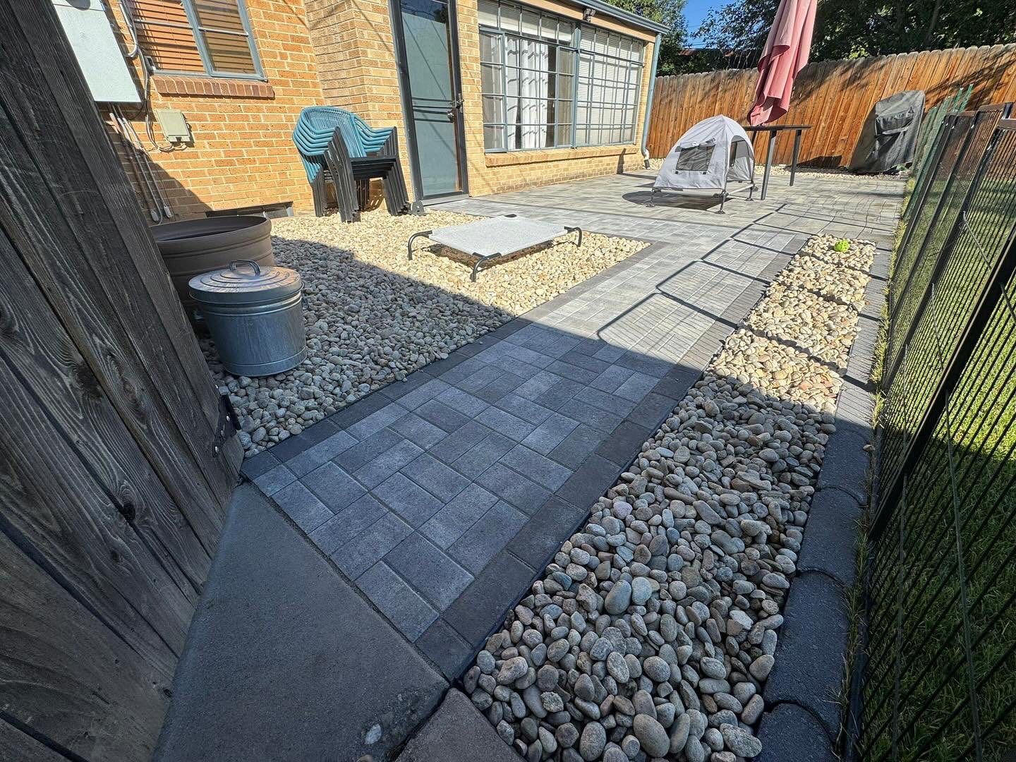 Stone patio with brick path, bordered by rocks, leading to a yard with a building.