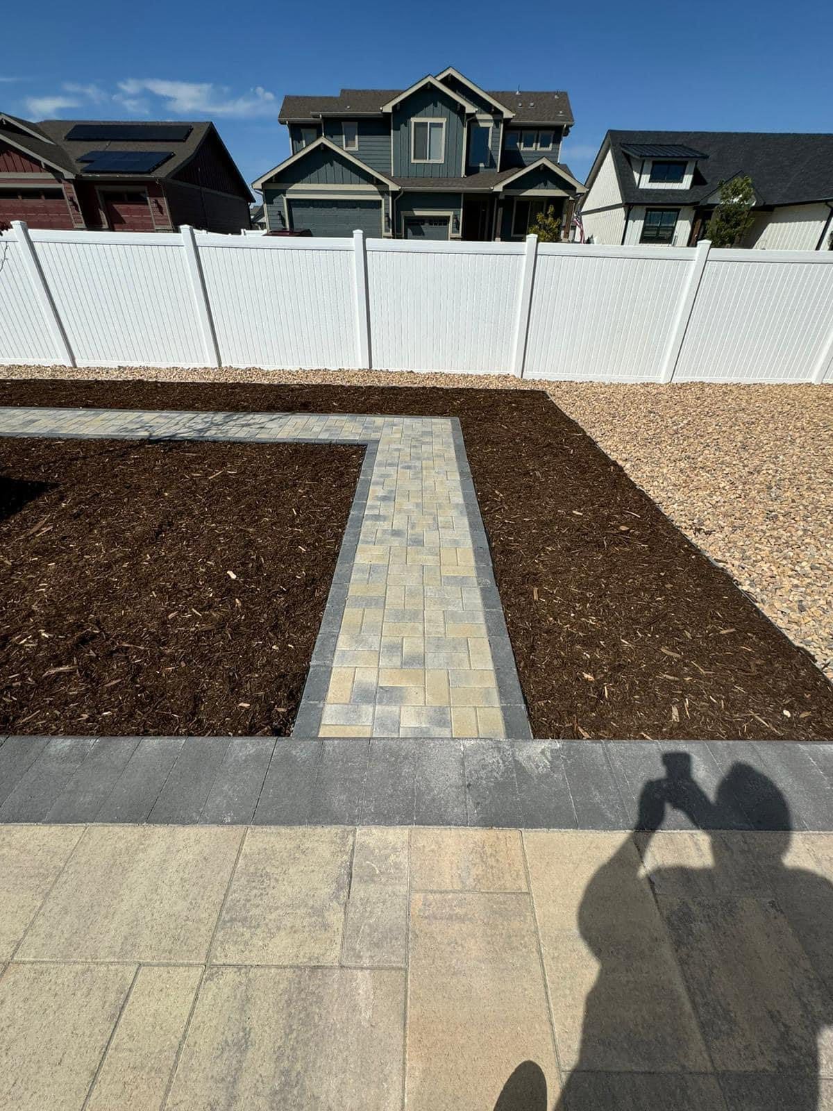 Brick walkway, mulch, and gravel border a residential property with a white fence and blue house.