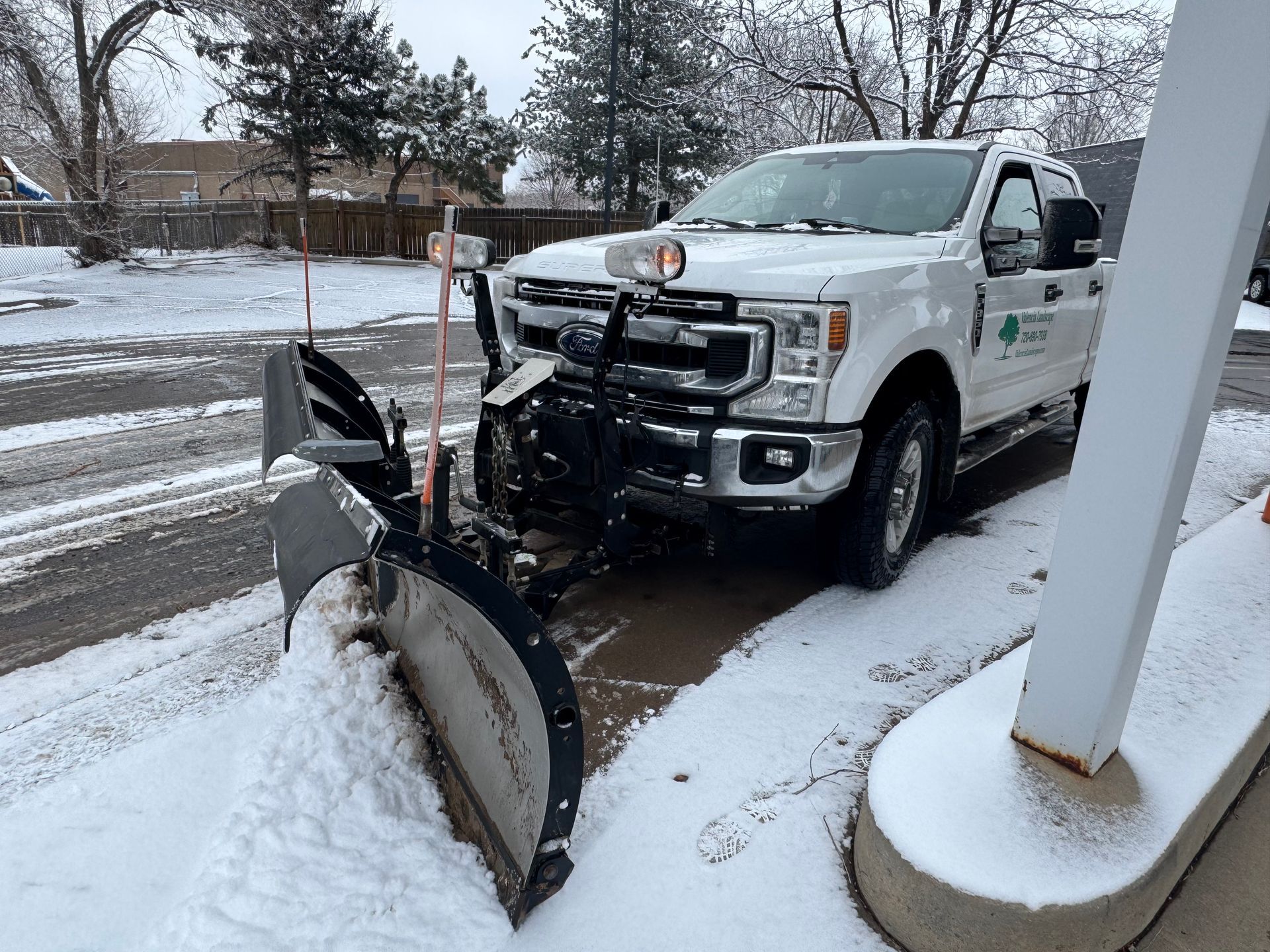 White snowplow truck clearing snow from a driveway, trees in the background.