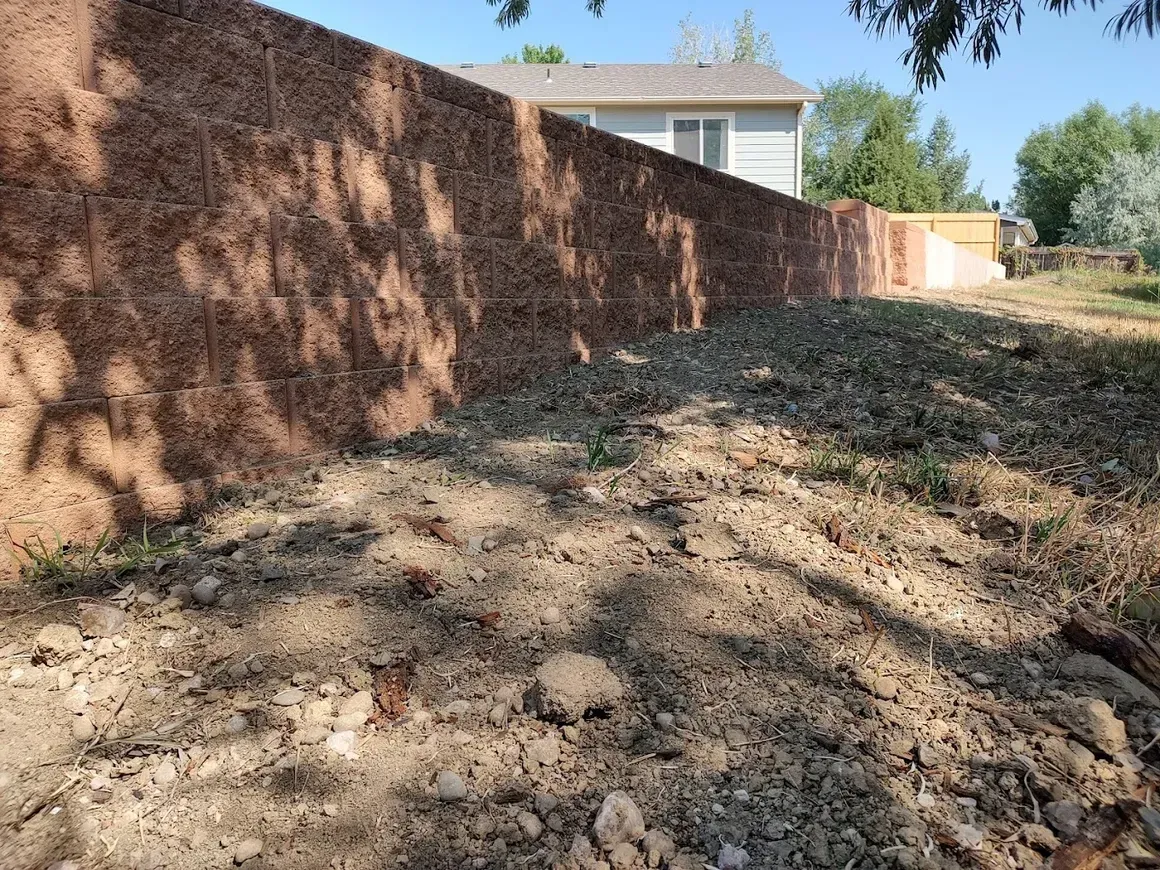 A dirt yard with a reddish retaining wall, house in the background.