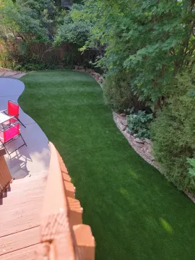 Artificial turf lawn bordered by stone and lush greenery, next to a deck with chairs.