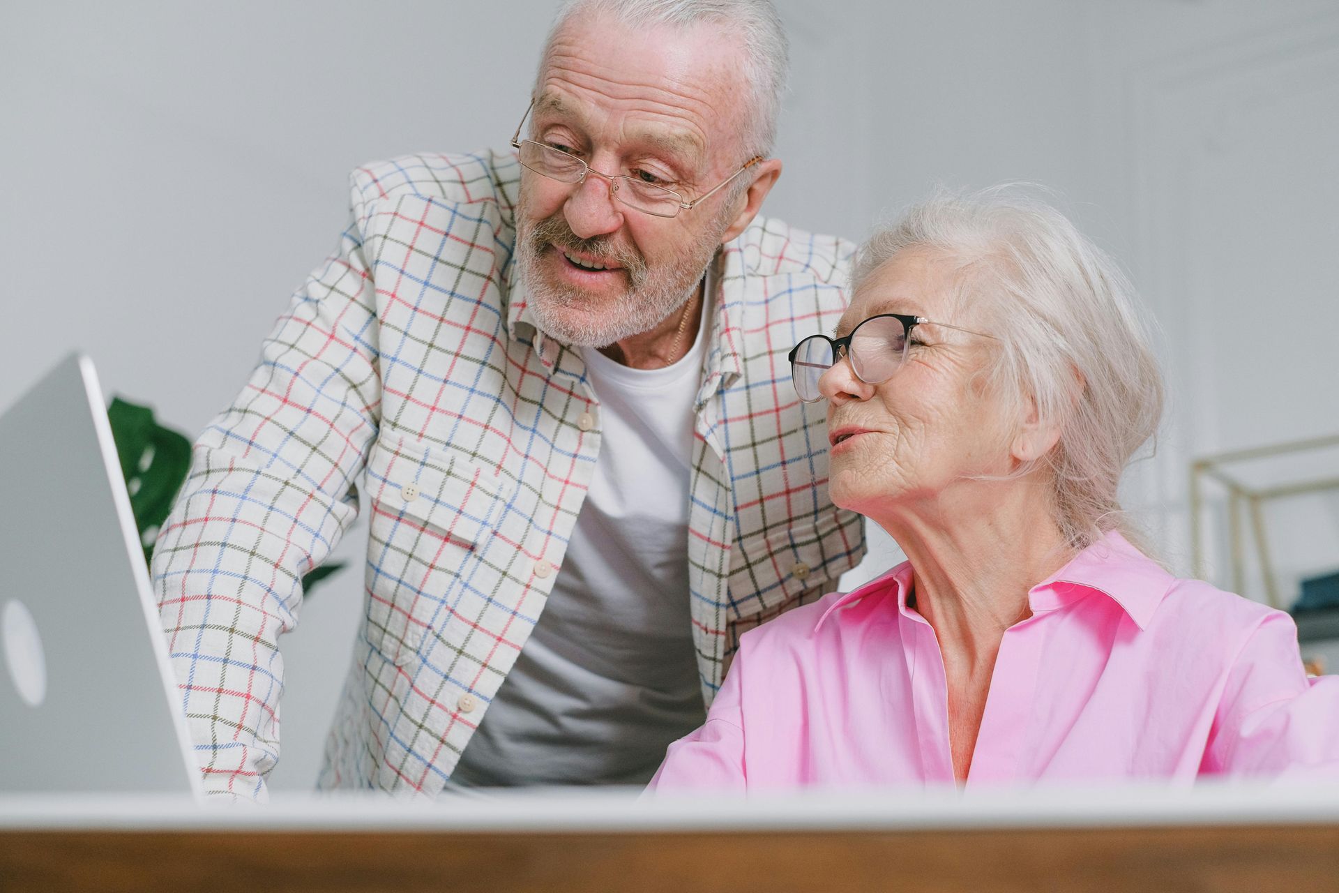 Two seniors smiling at a laptop, with the woman in a pink shirt and the man in a plaid jacket