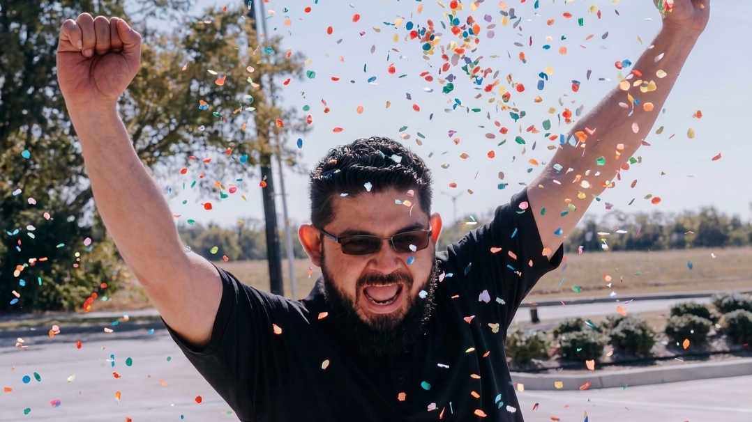 Man with raised arms celebrating amid falling confetti outdoors, wearing sunglasses and a black shirt.