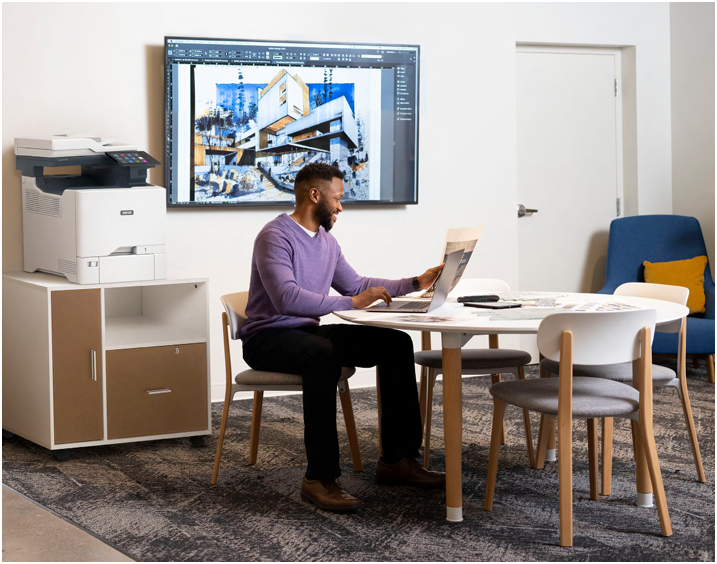 Person working at a table in a modern office, with a wall-mounted screen and printer nearby.