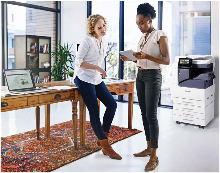 Two coworkers talking in a bright office beside a wooden desk and a printer