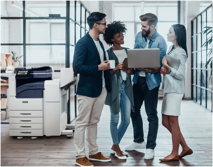 Four coworkers discussing a laptop in a bright office beside a printer and glass walls