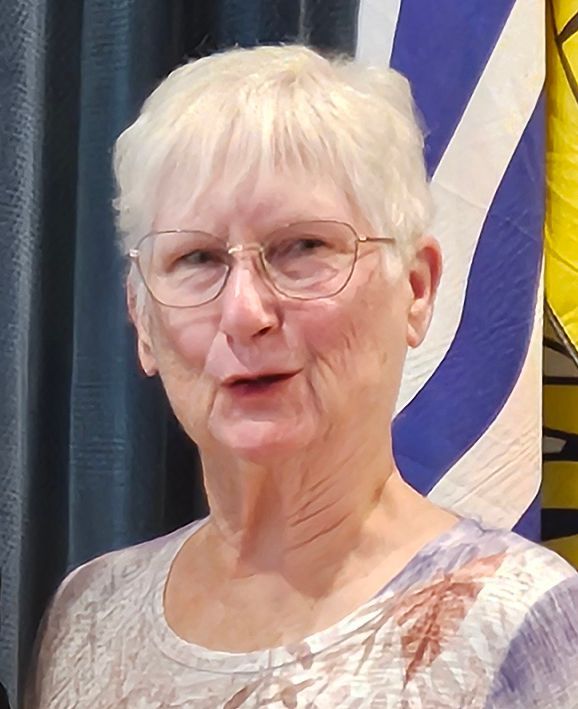 Woman with short white hair, glasses, and a patterned shirt smiles in front of a blue and white striped flag.