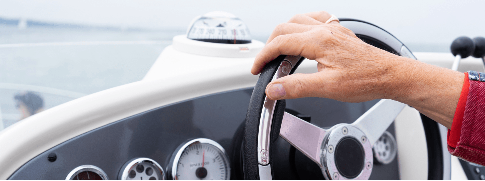 Hand gripping the steering wheel of a boat, with gauges and a compass in view.