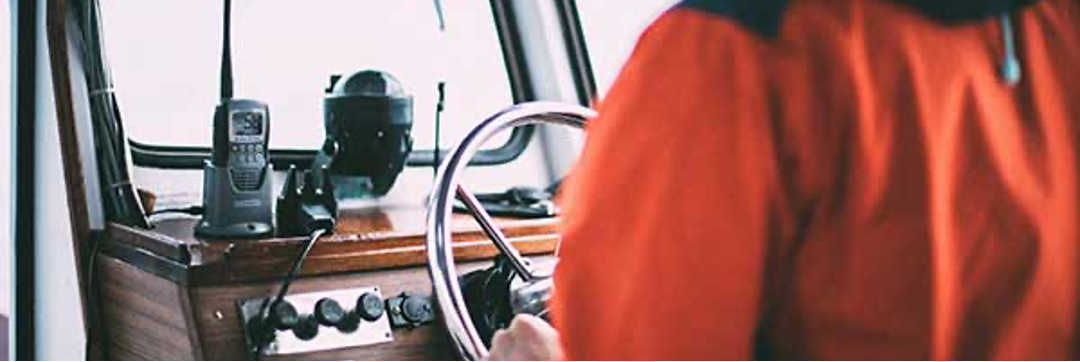 Person in red jacket steers a boat. Control panel, radio, and steering wheel are visible. Setting is outdoors, possibly on the water.