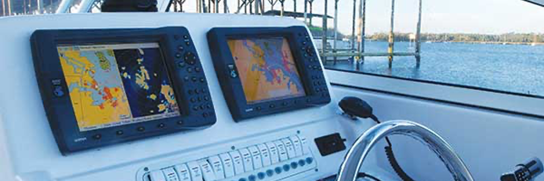 Boat's dashboard with two screens displaying navigation data. Steering wheel visible, with water and sky in the background.