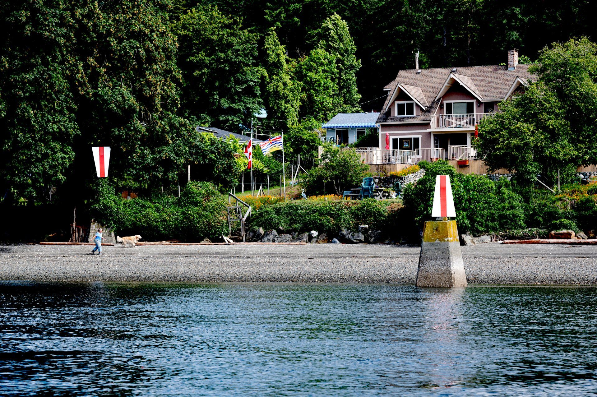 Beach scene with a house, trees, and buoys in the water. A person and dog are walking on the beach.
