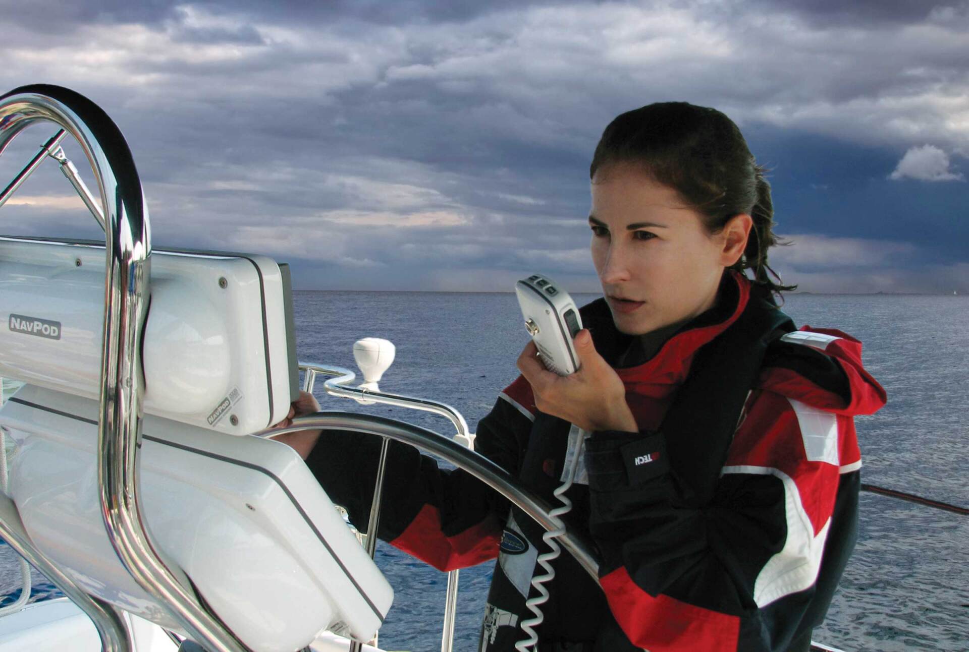 Woman on a sailboat speaking into a radio, holding the helm, with ocean and cloudy sky in the background.