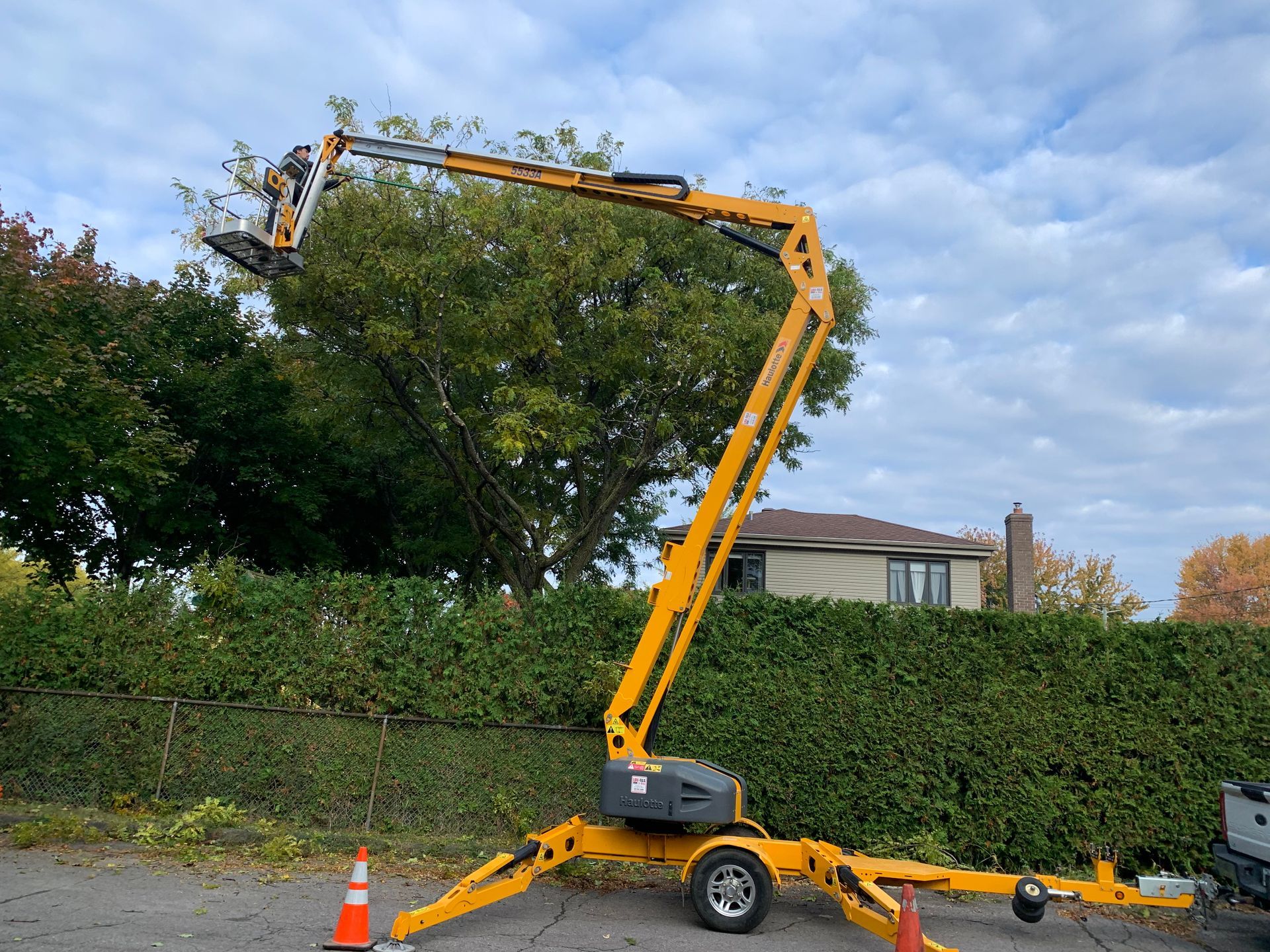 Une nacelle élévatrice jaune taille un arbre près d'une haie verte et d'une maison sous un ciel nuageux.