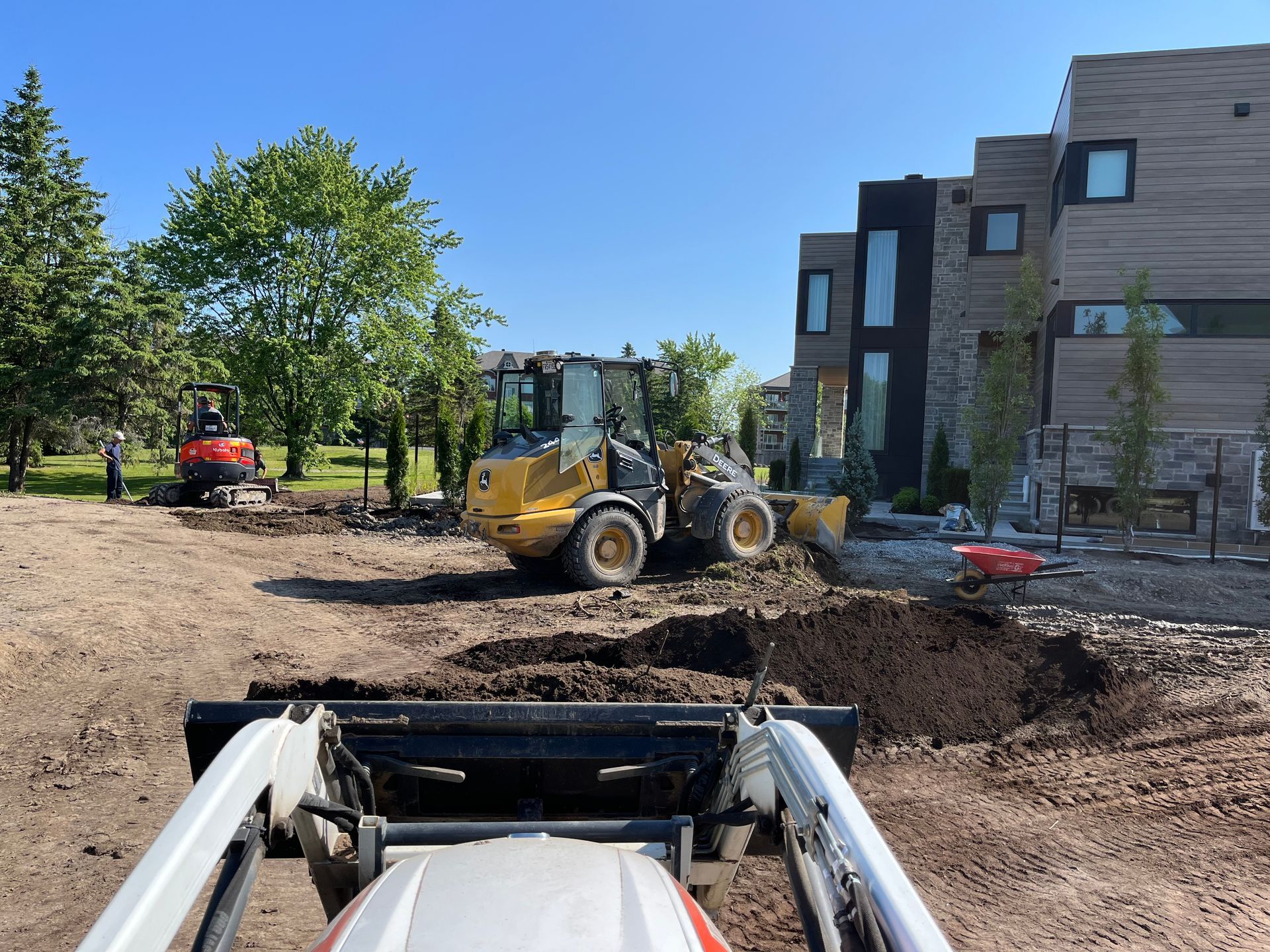 Chantier de construction avec une chargeuse compacte au premier plan et des engins de chantier près d'une maison moderne par une journée ensoleillée.