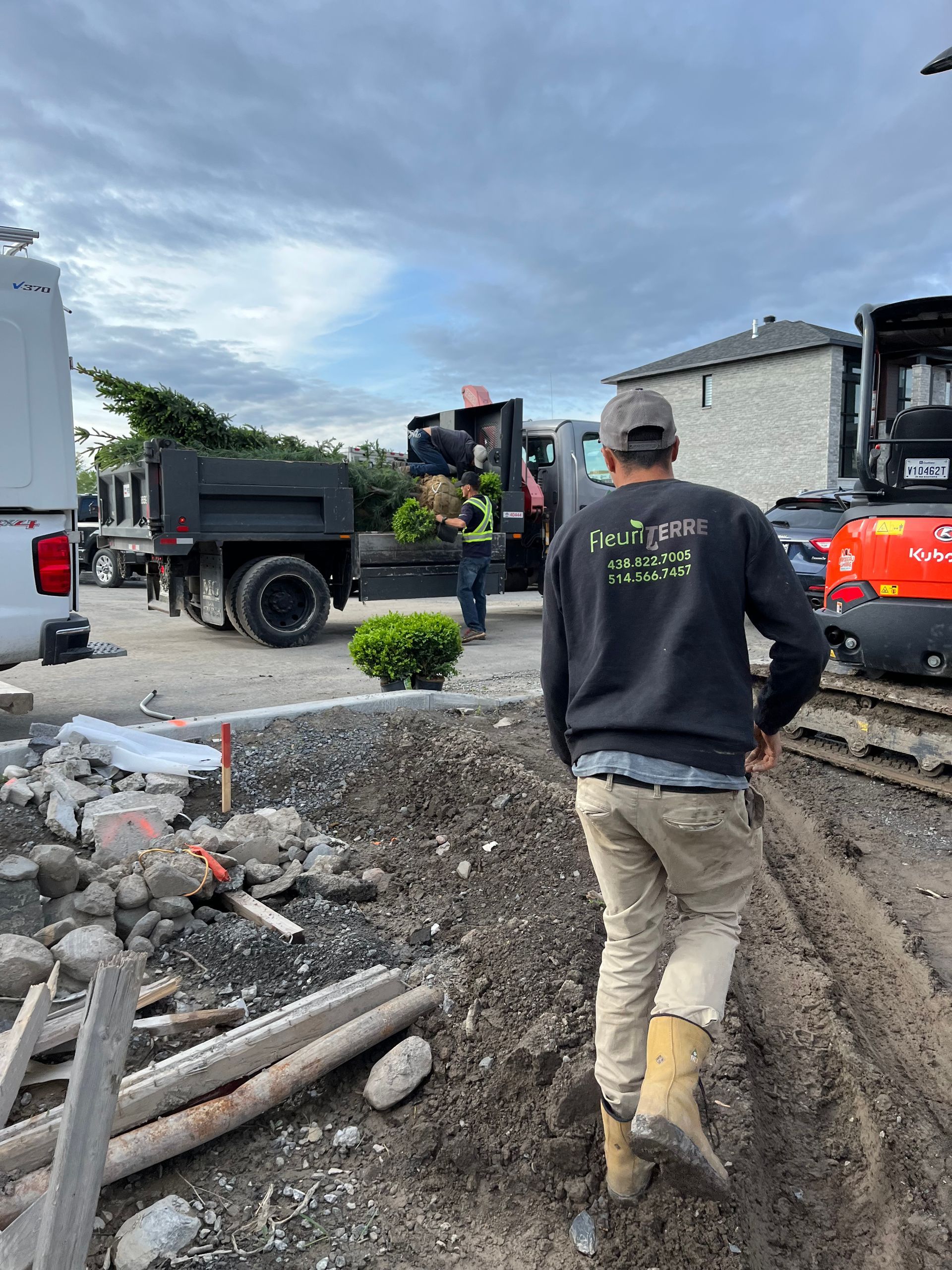 Un homme en tenue de travail se dirige vers un camion-benne sur un chantier. D'autres ouvriers et des engins de chantier sont présents.