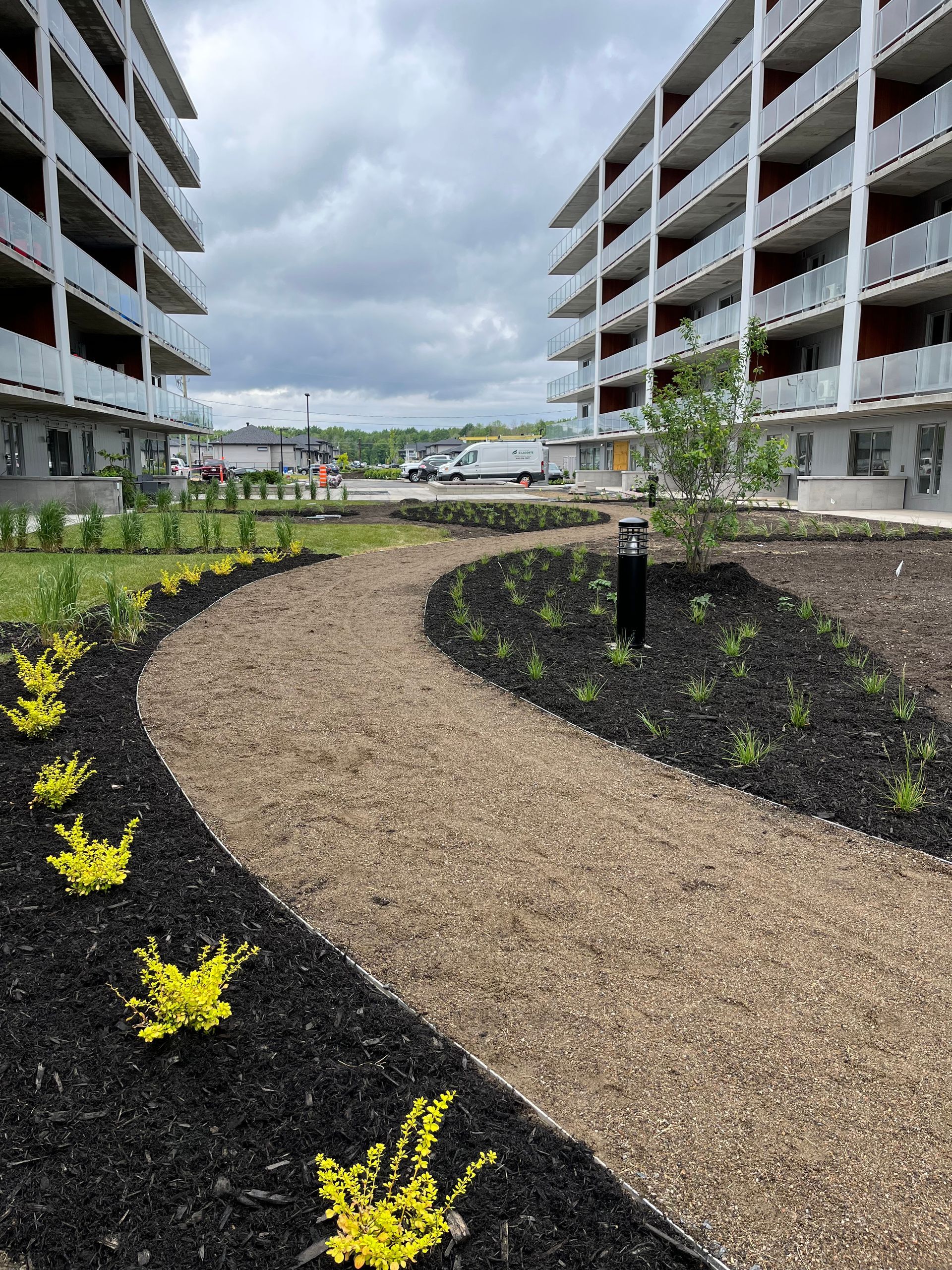 Allée recouverte de copeaux de bois et agrémentée d'aménagements paysagers entre des immeubles d'appartements par temps nuageux.