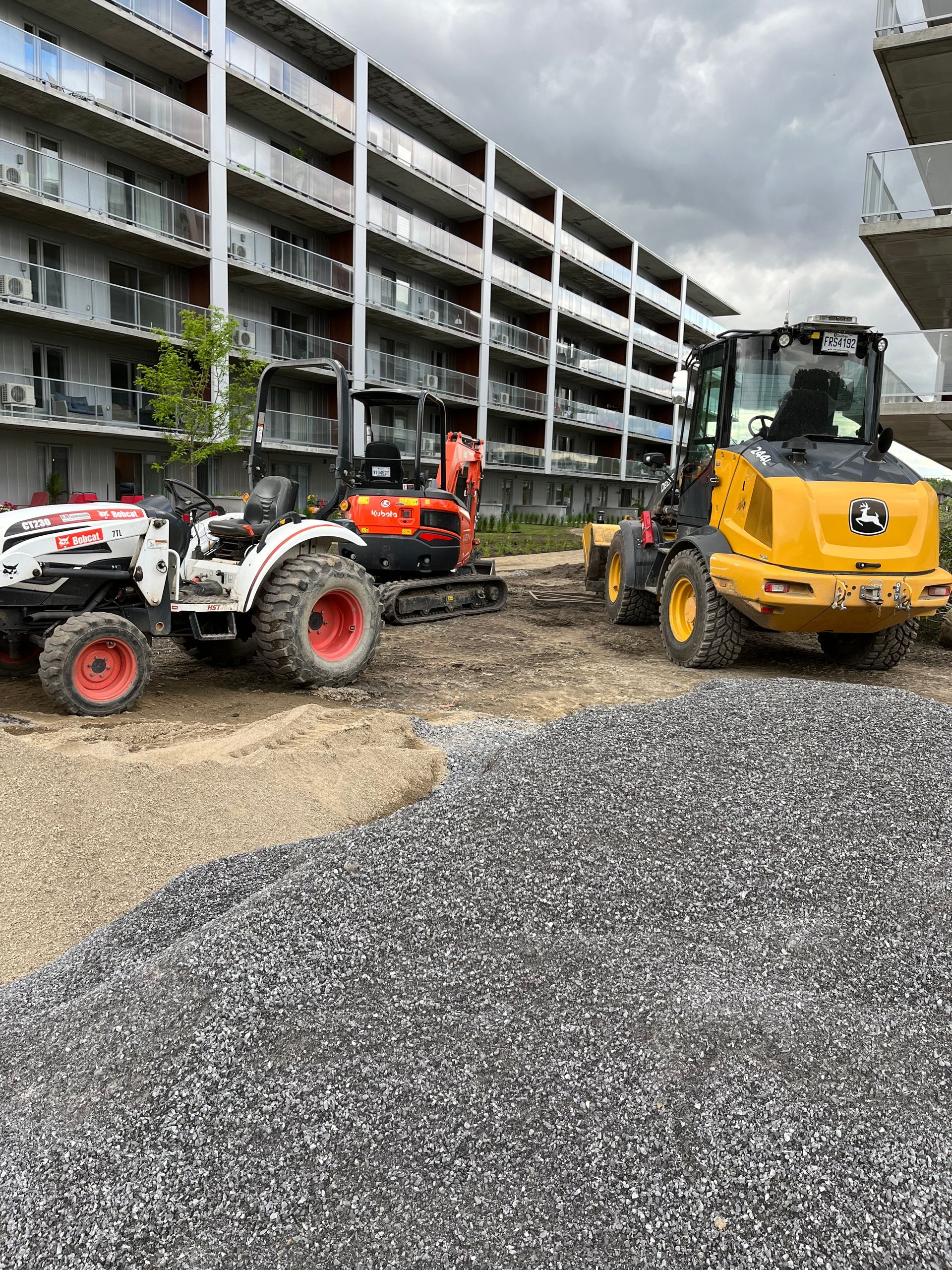 Chantier de construction avec du gravier, une chargeuse John Deere et un tracteur avec pelle mécanique, près d'un immeuble d'appartements.