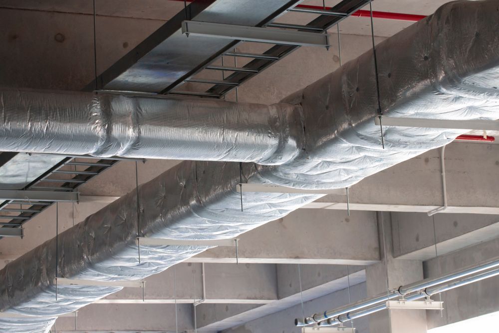 Insulated HVAC ductwork, silver in color, runs across a concrete ceiling in a construction setting.