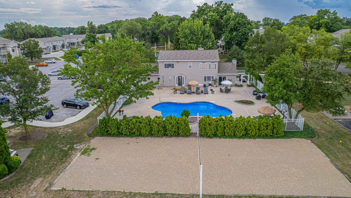 Backyard with pool, volleyball court, and house. Green trees surround a blue pool; a beige volleyball court is in front.