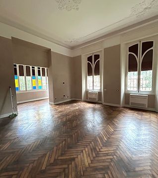 Elegante stanza vuota con pavimento in legno a spina di pesce, alte finestre ad arco e pareti color beige tenue.