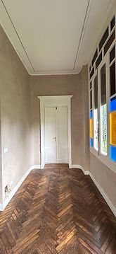 Stretto corridoio con pavimento in legno a spina di pesce, pareti beige, porta bianca e finestre colorate sulla destra.