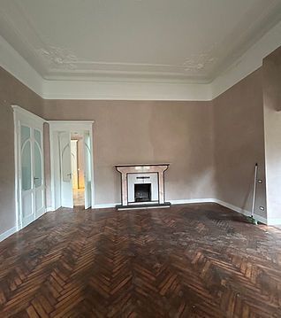 Soggiorno vuoto con pavimento in legno a spina di pesce, soffitto bianco decorato e un piccolo camino sulla parete di fondo.