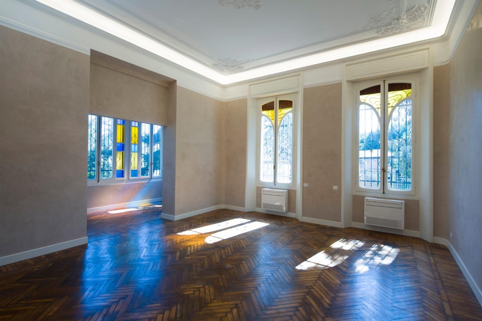 Stanza vuota e luminosa con pavimento in legno, finestre alte e pareti beige.