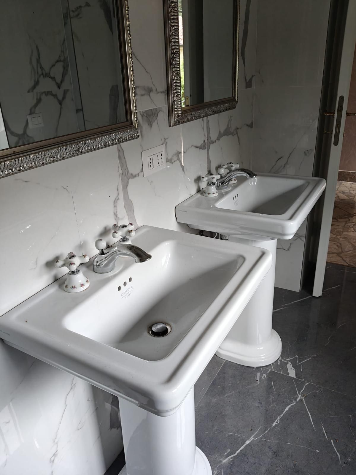 Due lavabi bianchi a colonna in un bagno piastrellato, con specchi sopra e crepe visibili nel muro.