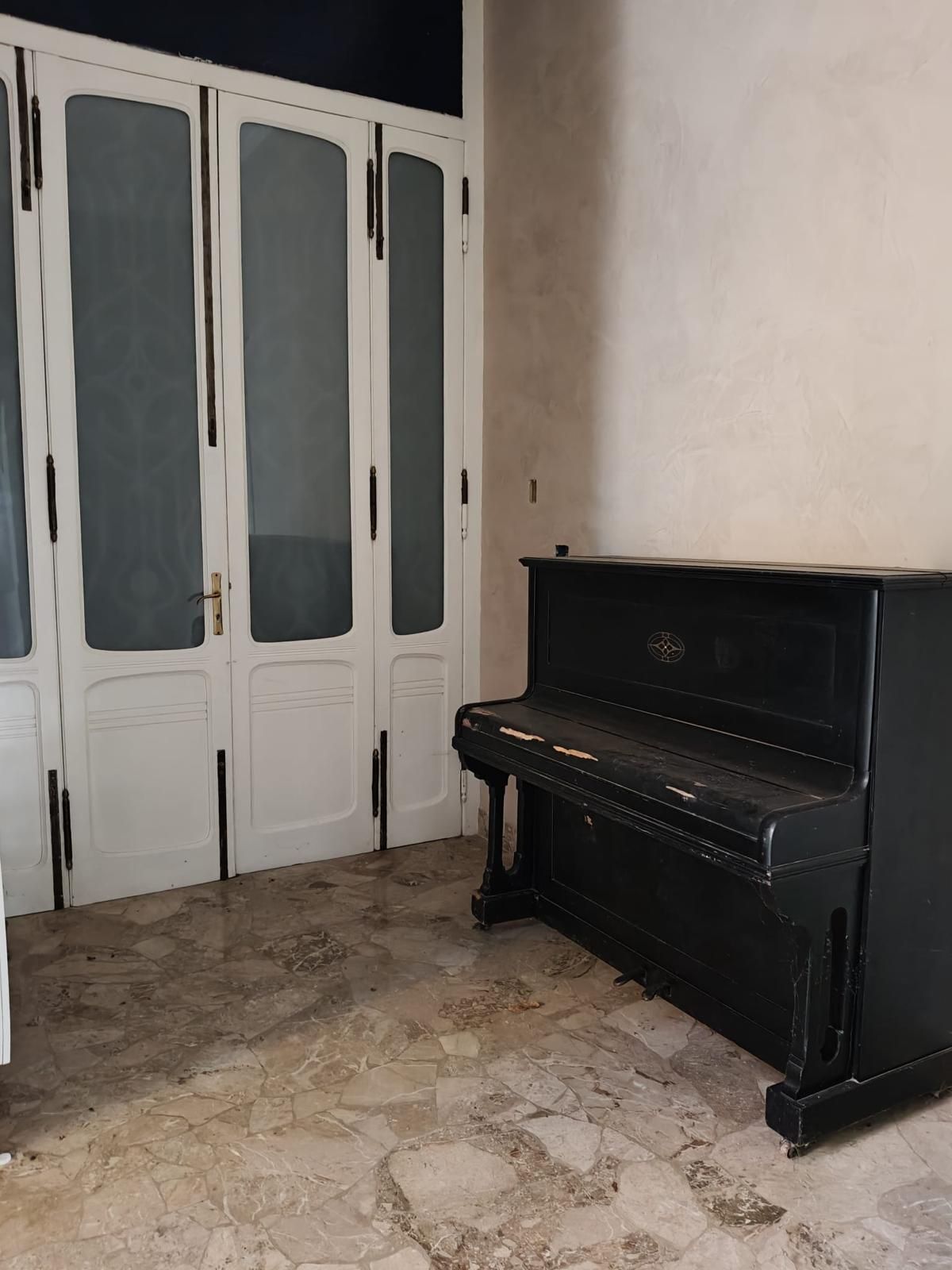 Stanza vuota con armadi bianchi sulla sinistra e un pianoforte verticale nero contro una parete beige.