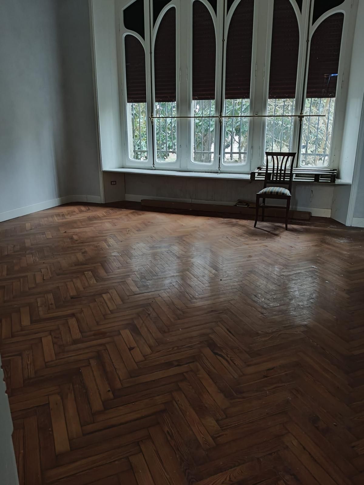 Stanza vuota con pavimento in legno a spina di pesce, alte finestre ad arco e una sola sedia accanto alla finestra.