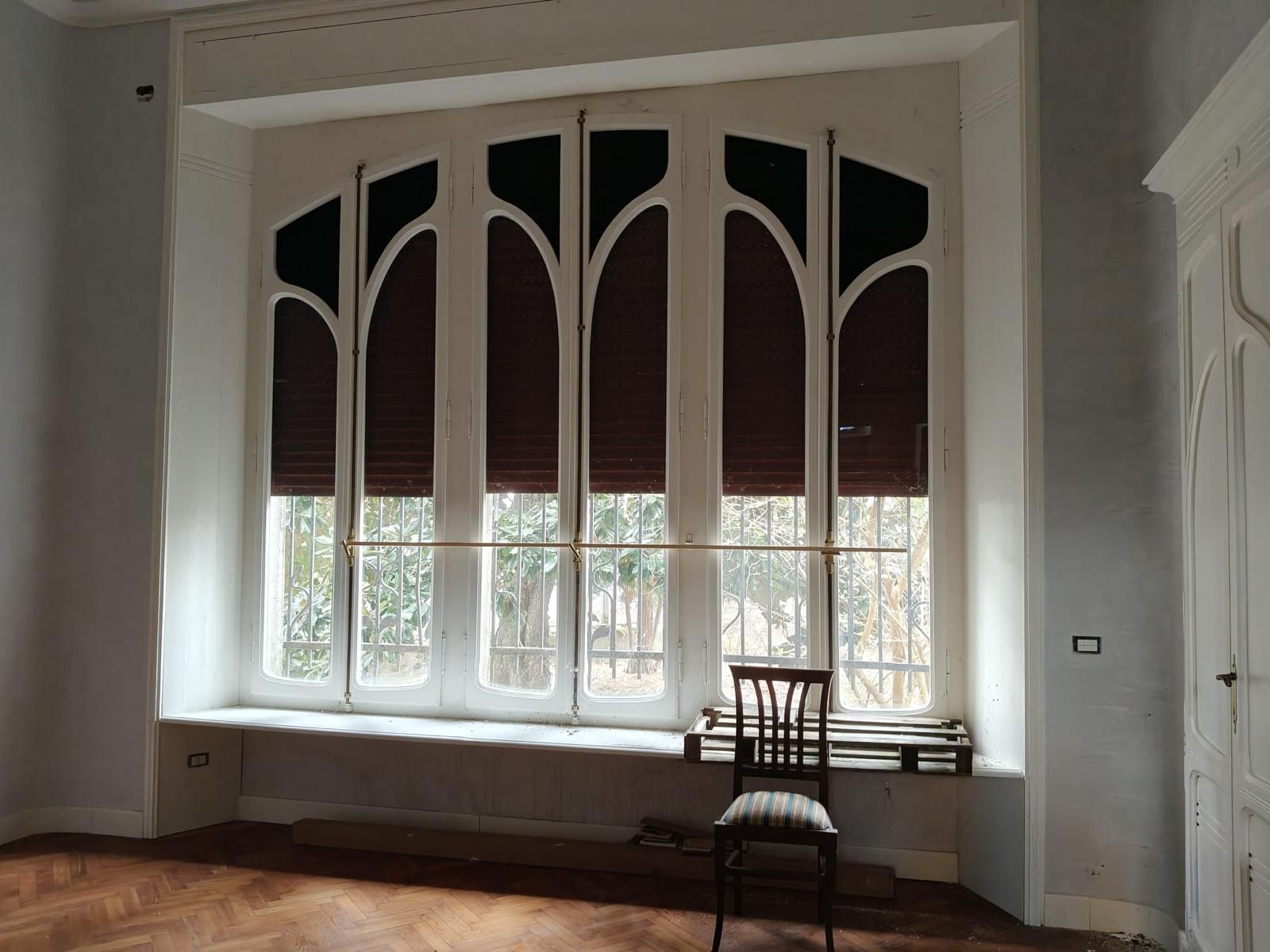 Stanza vuota con una grande finestra ad arco, pareti bianche e due sedie di legno sul davanzale.