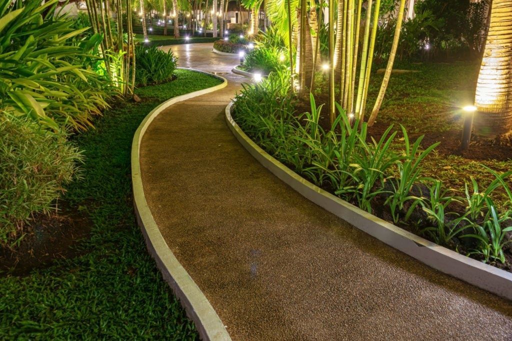 Curving gravel path at night, lit by small ground lights, bordered by grass and greenery.