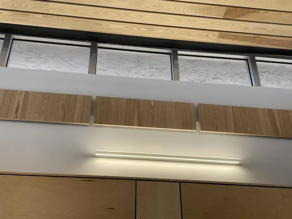 Wooden panels with a strip light on a white wall, above a glass and wood ceiling.