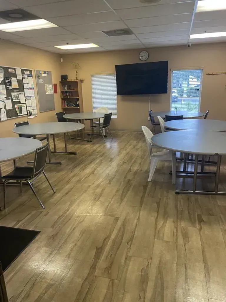 An empty community room with round tables, chairs, a TV, and a corkboard. Wooden floor, beige walls.