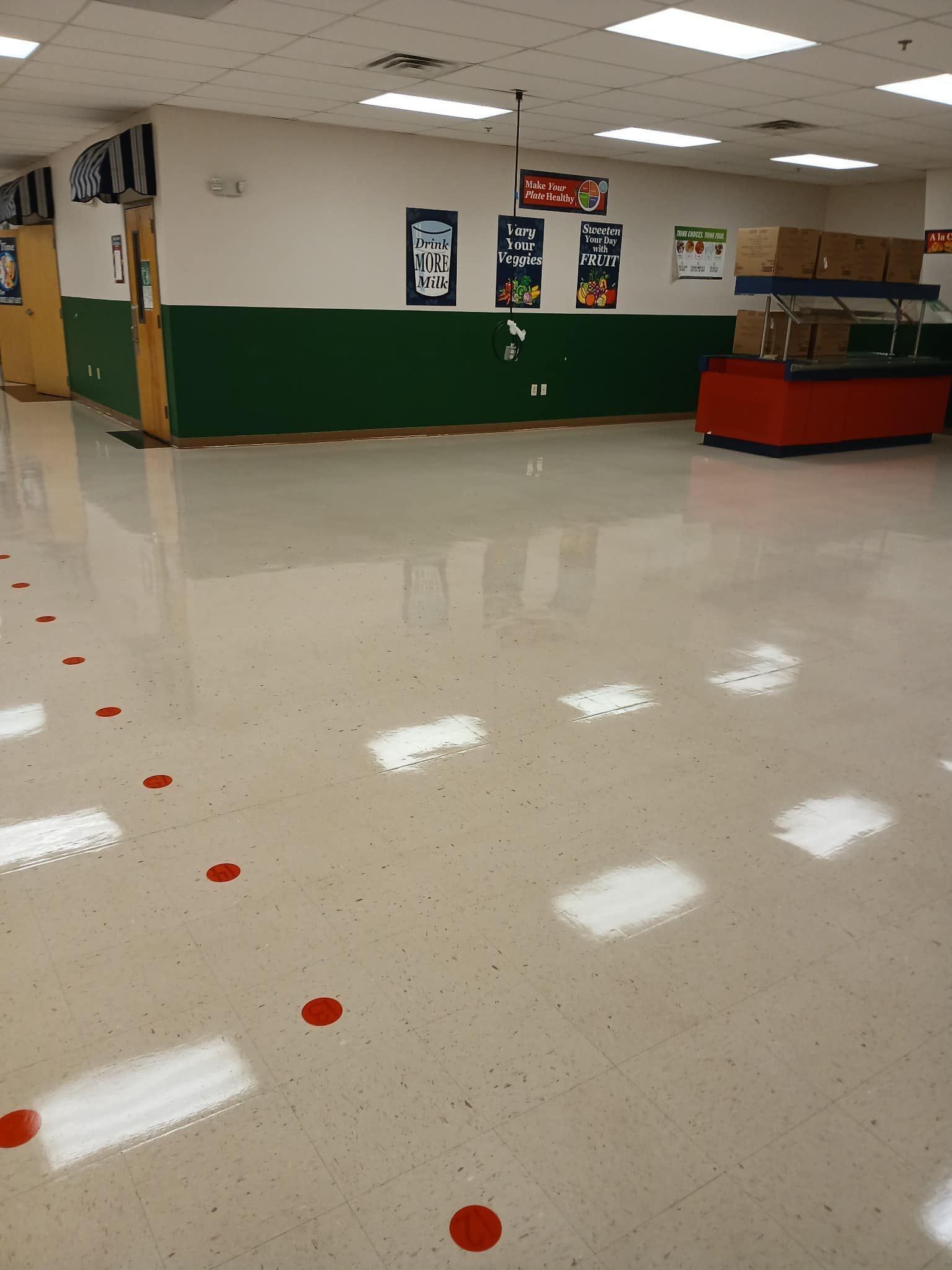 Empty cafeteria with glossy floor, green and white walls, food serving station in the background, and red dots on the floor.