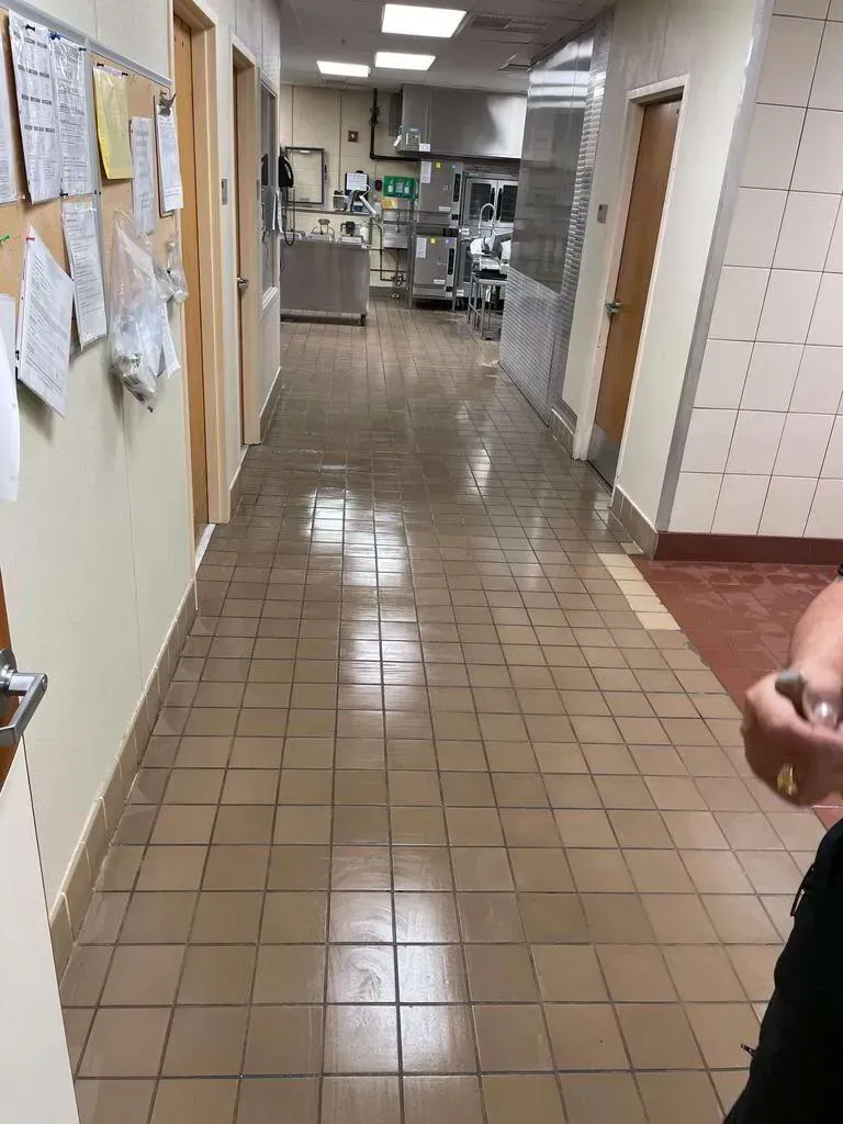 Long, tiled hallway in a commercial kitchen with doors, bulletin board, and equipment visible in the distance.
