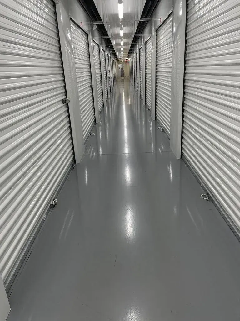 Long hallway with rows of closed storage unit doors, a shiny gray floor, and bright overhead lights.