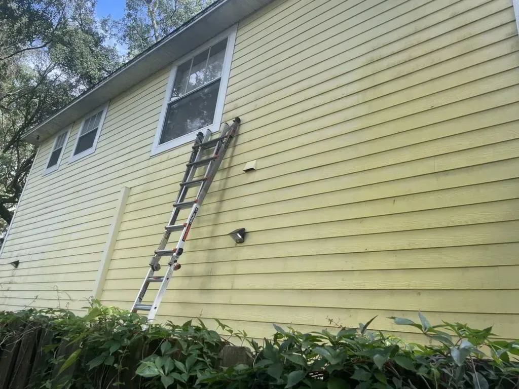 Ladder leans against a two-story yellow house with greenery in the foreground.