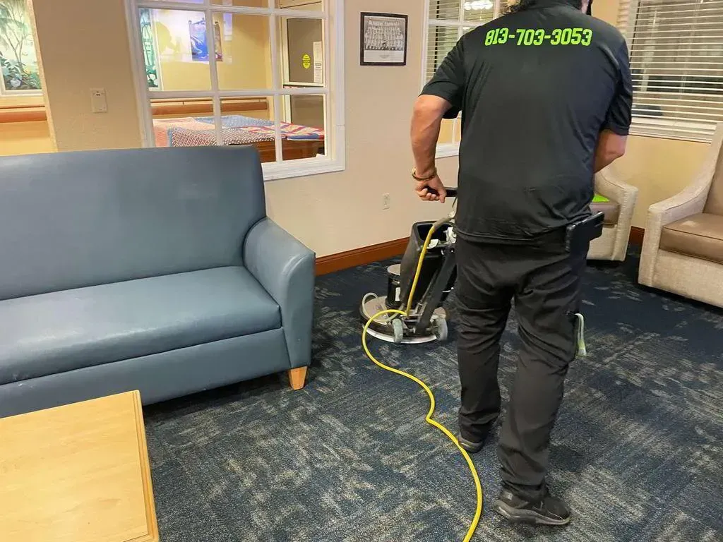 Person in black uniform cleaning carpet with a machine, near a blue couch and white chairs.